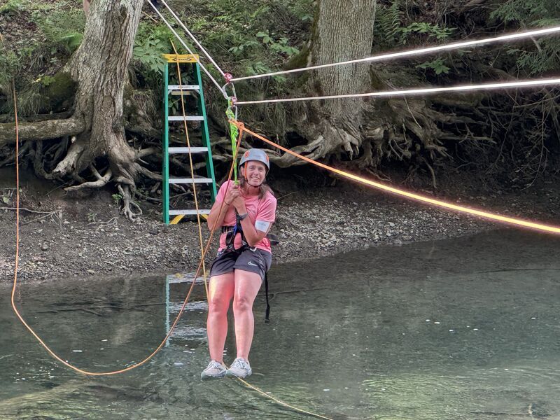 The image shows a person on a zip line over water. They are wearing a helmet and safety gear. There is a ladder in the background. The person appears to be smiling and enjoying the activity. The setting seems to be a natural environment with trees and water.